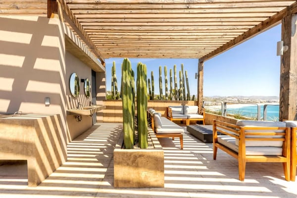 Pergola-covered terrace with wooden furniture, tall cacti, and turquoise sea view