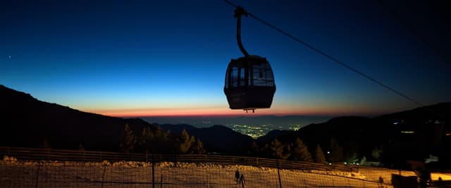 Silhouetted cable car suspended above mountain valley at dusk with city lights below