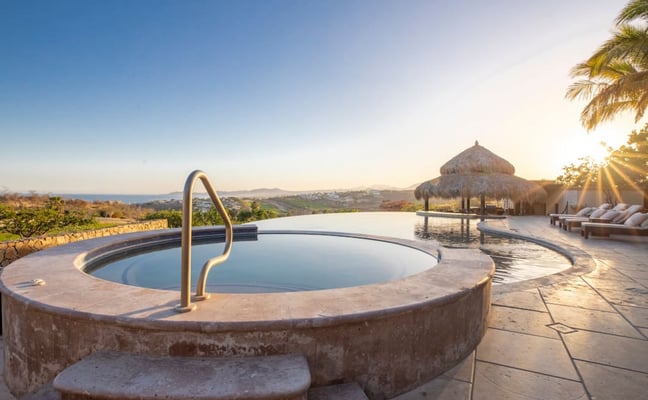 Circular hot tub with metal handrail on terrace overlooking infinity pool, thatched cabana, and coastal landscape at sunset