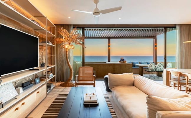 Open-plan living room opening onto a beachfront terrace, with a large sofa, dining table, and ocean view
